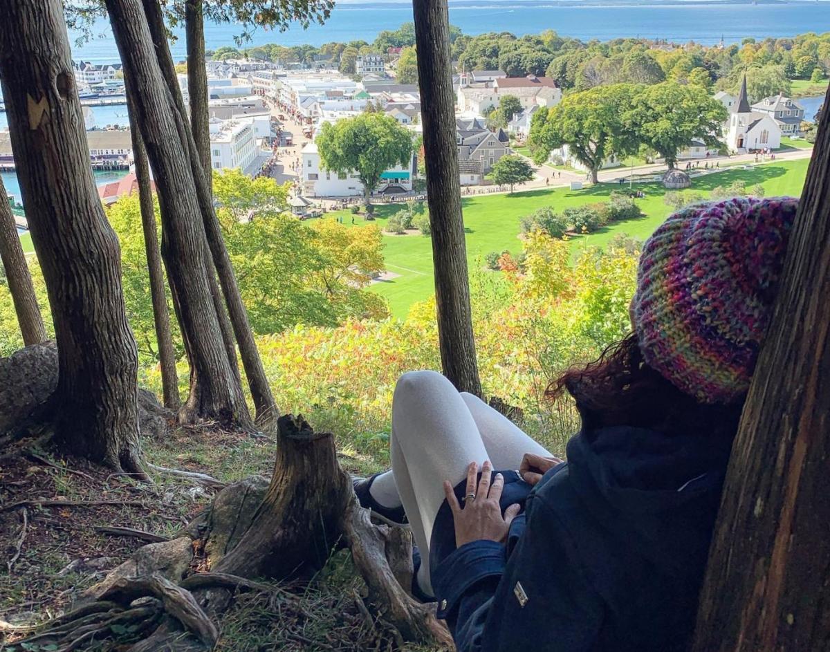 A woman wearing a beanie cap sits in the shade of a tree on a high bluff overlooking downtown Mackinac Island