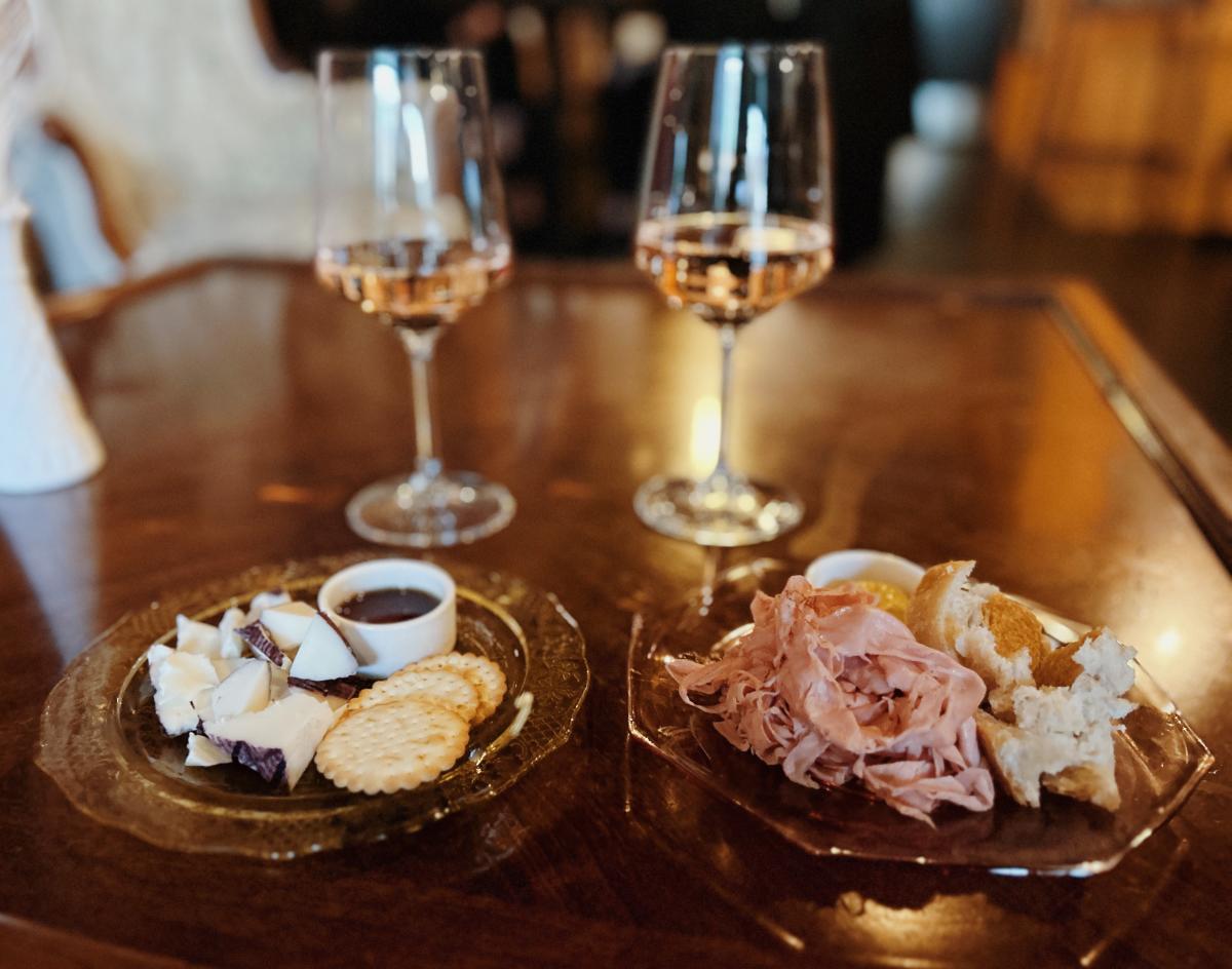 Two glasses of rosé wine on a wooden table flanked by plates of cheese, crackers and prosciutto with bread. The ambiance is warm and inviting.