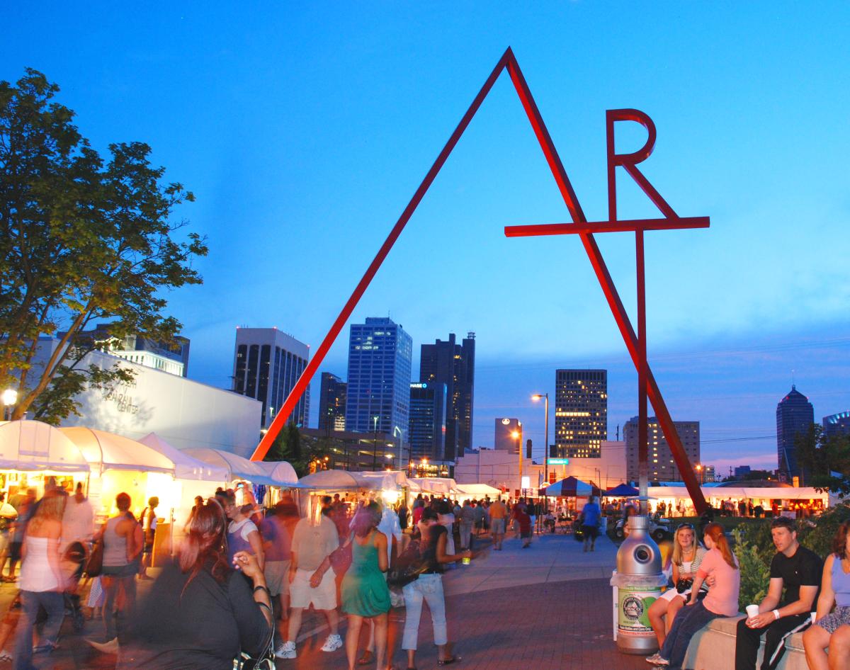 Buzzing crowd of patrons passing vendor booths in street under "ART" sign at Columbus Arts Festival