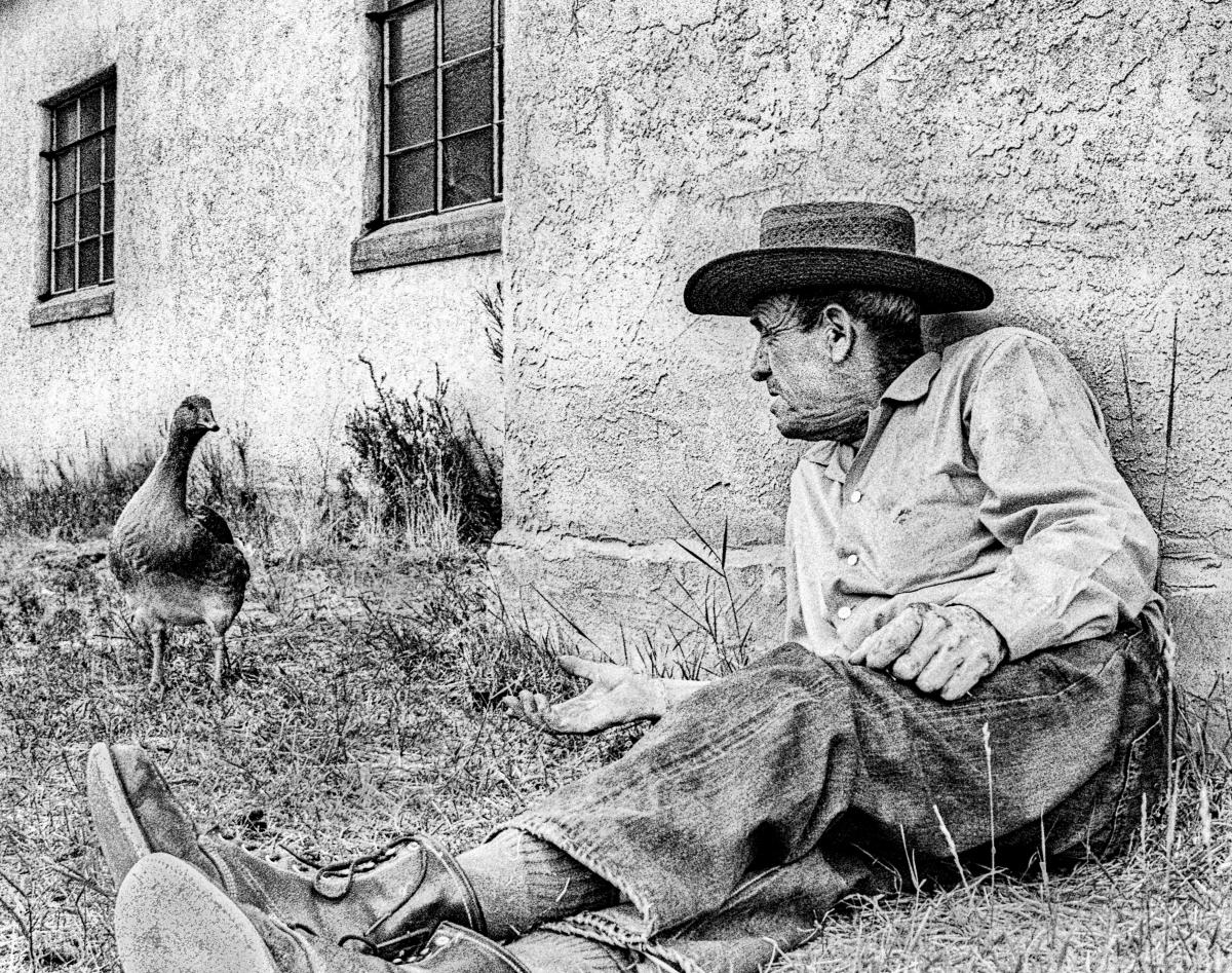 Black and white photograph of a man wearing a wide-brimmed hat sitting against an adobe wall outdoors.