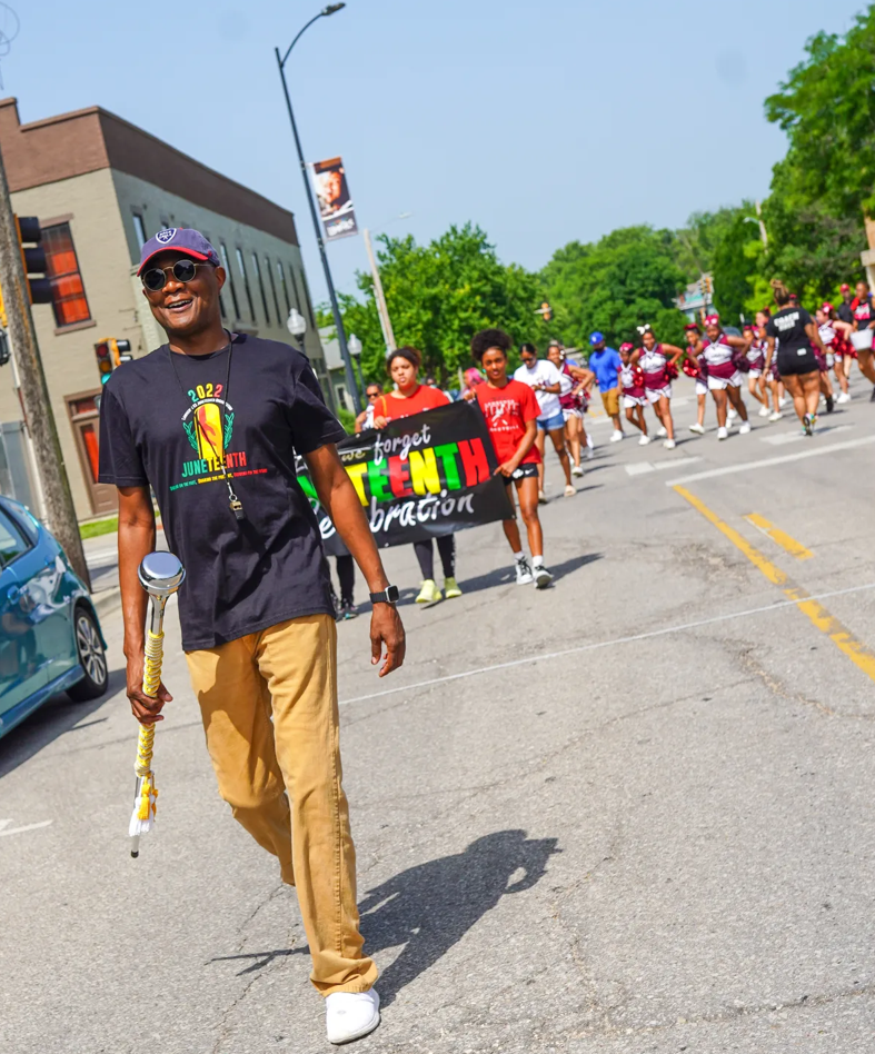 Photo taken at the Lawrence Juneteenth parade which features a man smiling in the foreground and cheerleaders in the background.