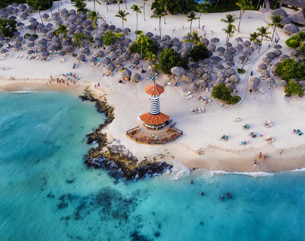 Aerial view of a tropical beach with white sand, palm trees, and a small lighthouse on a rocky outcropping. The water is turquoise and clear, with people swimming and sunbathing.