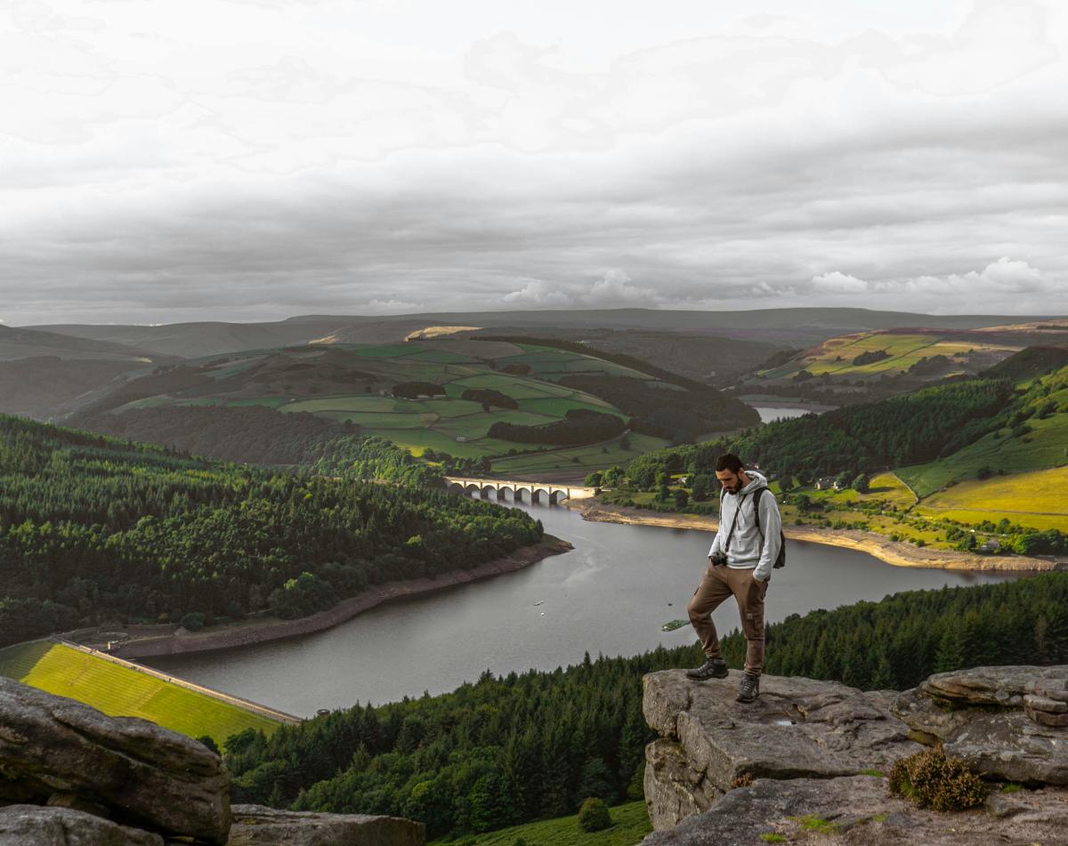 Person standing on the edge of a rocky cliff overlooking a lake in a wide valley, with green hills, forested slopes and winding water below under an overcast sky.