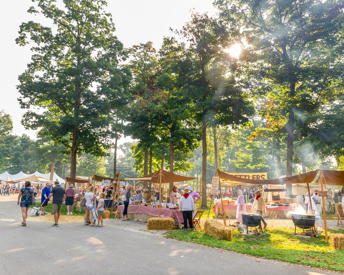 Food tents at the Johnny Appleseed Festival. People walking around looking at the food. Food vendors wearing pioneer-style clothing for the festival.