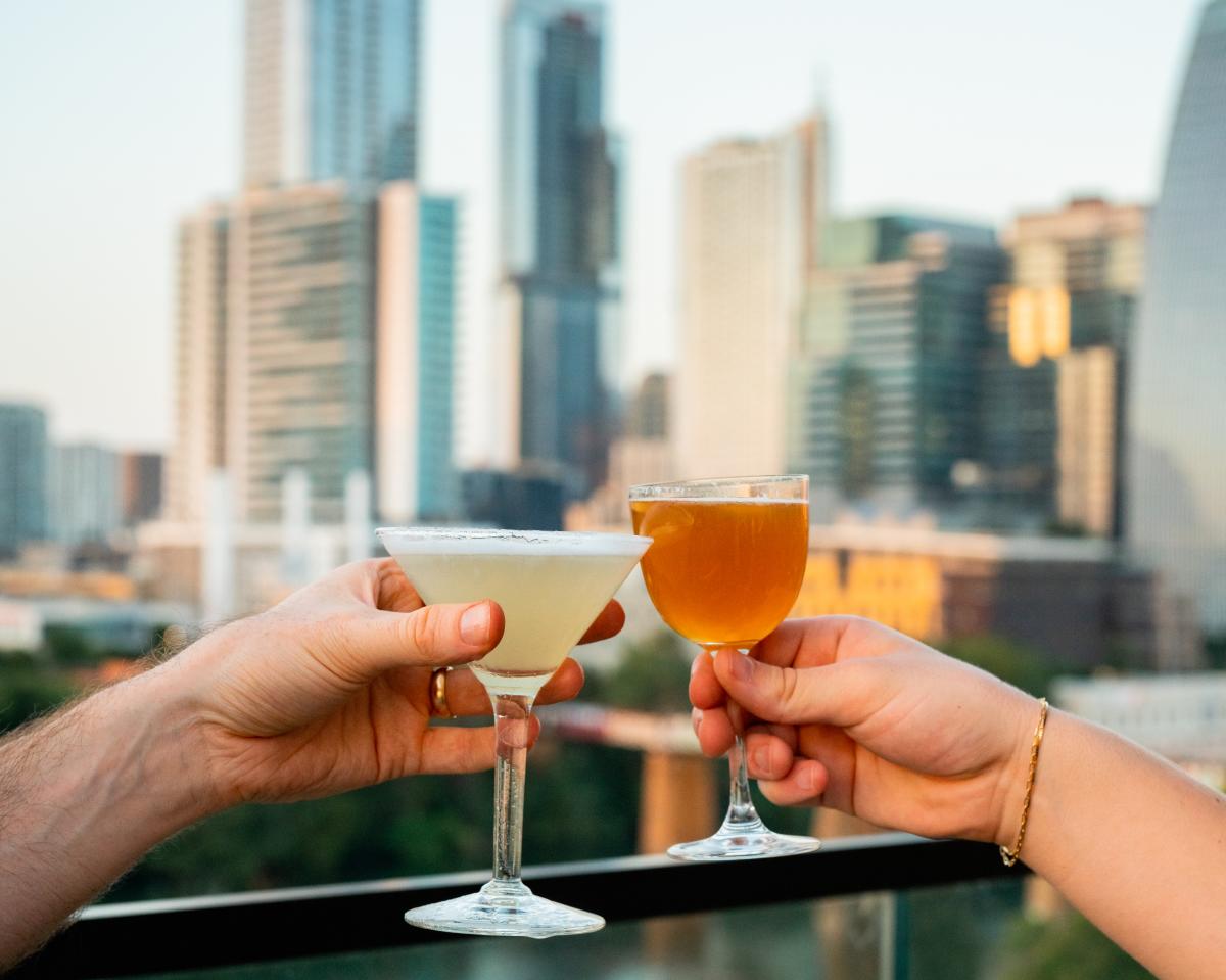 Image of two hands holding cocktails on the Nido rooftop with the Austin skyline in the background.