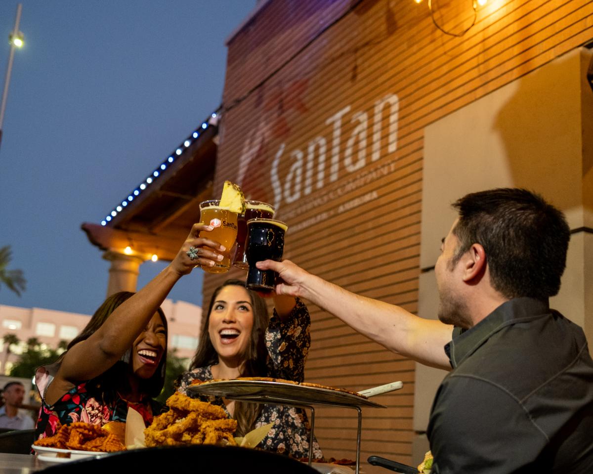 Two women and one man cheers beers on SanTan Brewing patio with food.