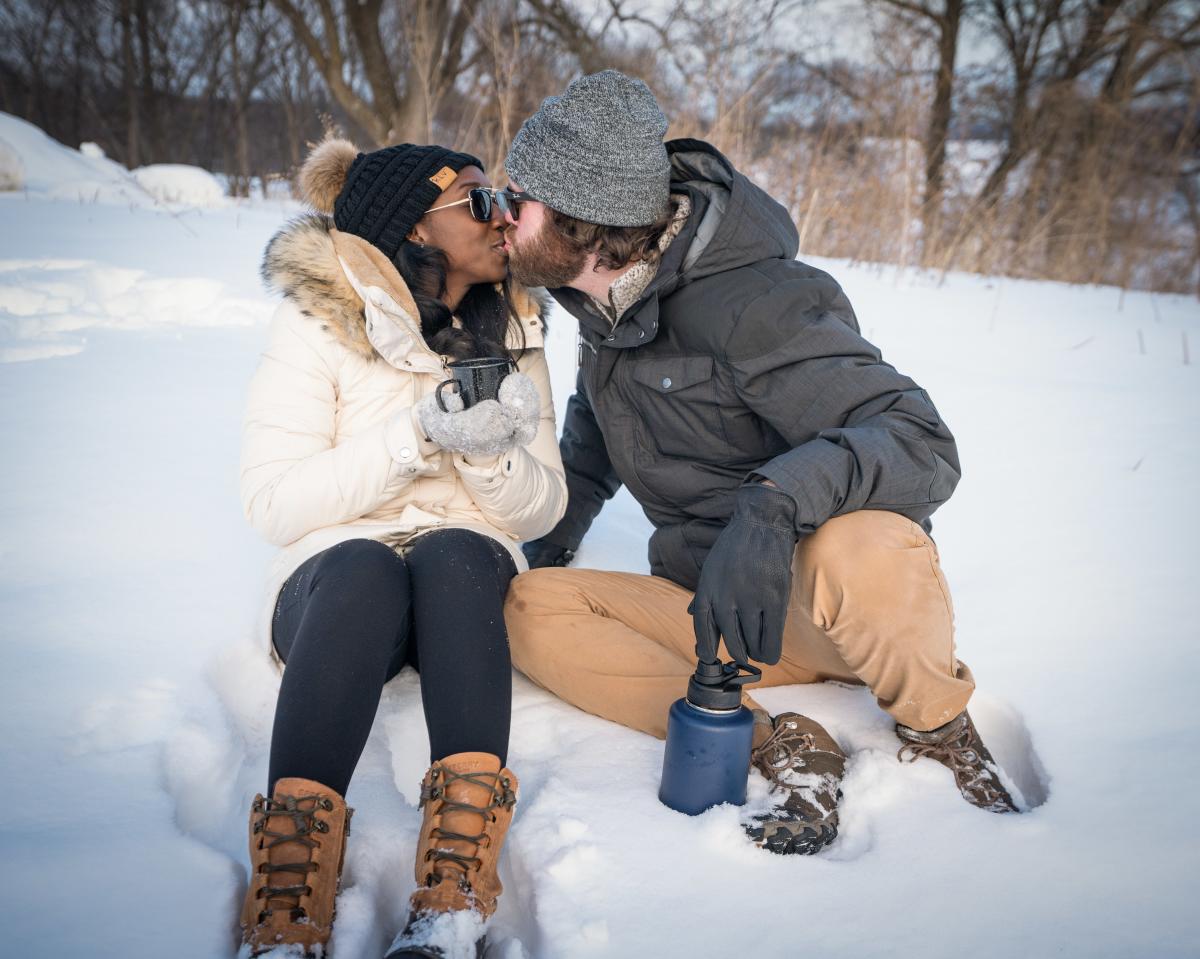 Two people sitting in snow, kissing. Wearing winter coats, boots, and hats.
