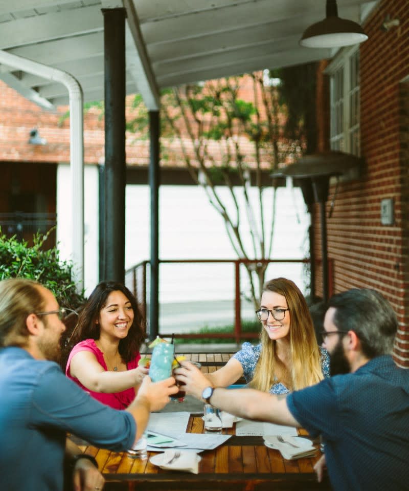 A group of people clink their cups together and smile while seated at a table on an outdoor patio.