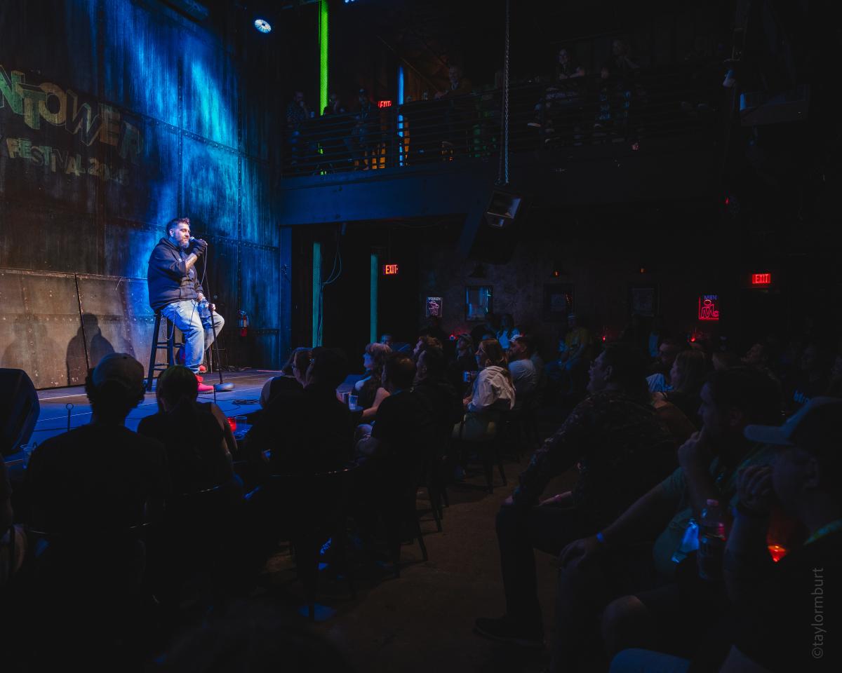 A man sitting on a stool onstage with a mic during a comedy set at Vulcan Gas Company.