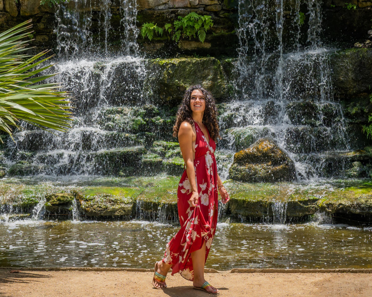 Photo of woman in red dress walking in front of waterfall and green trees at Austin Botanical Gardens