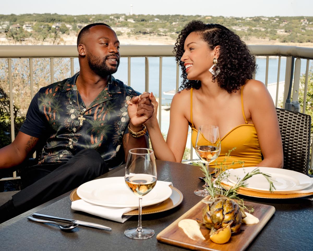 A couple sitting at an outdoor table holding hands with food and wine on the table.