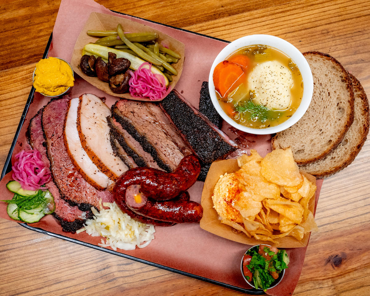 Flat lay image of a tray of smoked meats, side dishes and bread sitting on a tray on a table at Mum Foods.