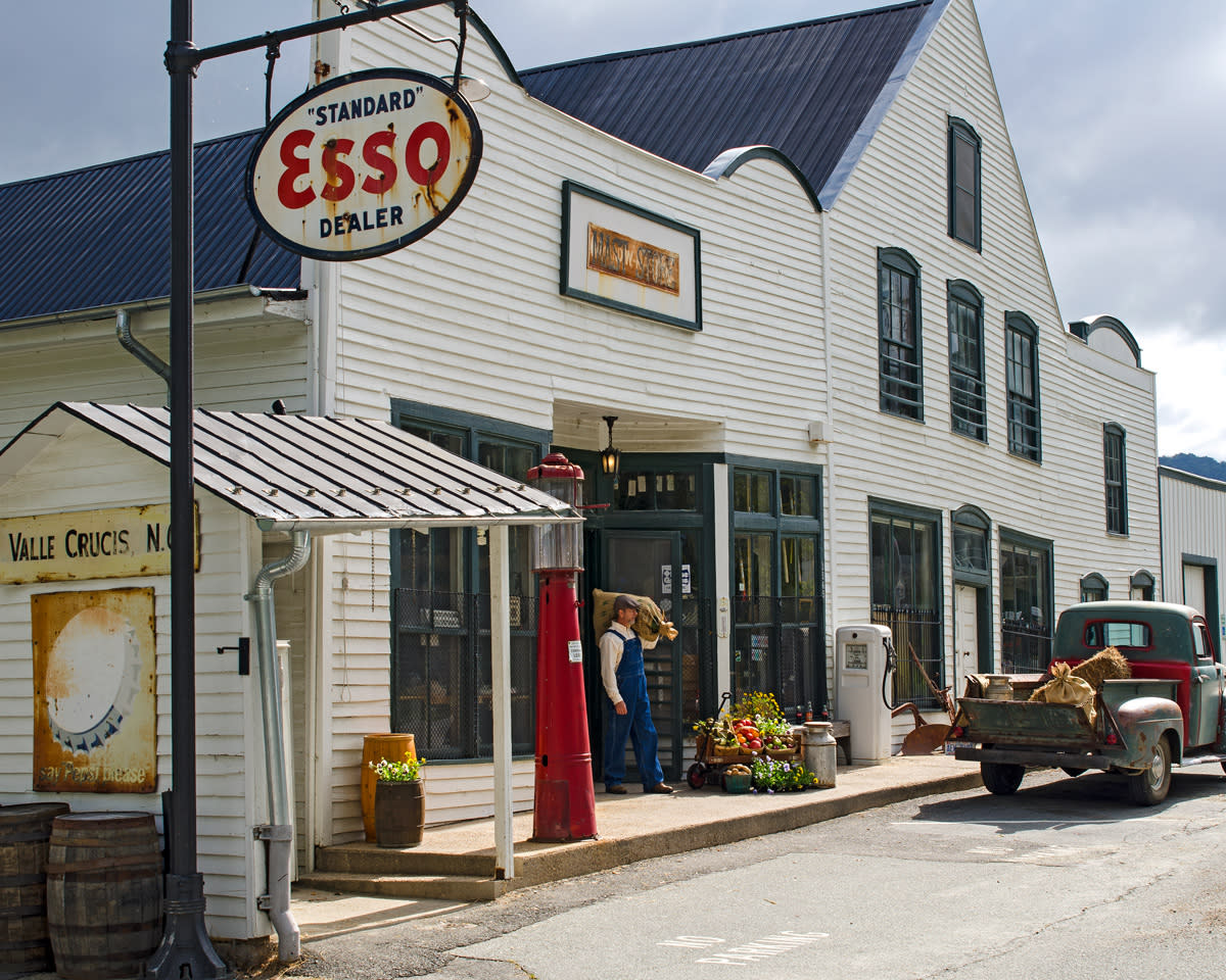 A nostalgic, white shiplap facade of the Original Mast General Store in Valle Crucis with a vintage Esso gas sign hanging in the foreground. In the doorway of the storefront, a man in overalls carries a large sack on his shoulder to an antique truck.