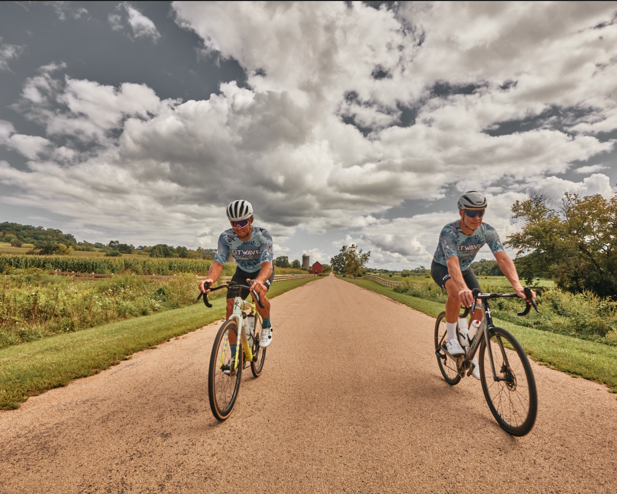 Two cyclists riding their bikes on a backroad, with a cloudy sky behind them.