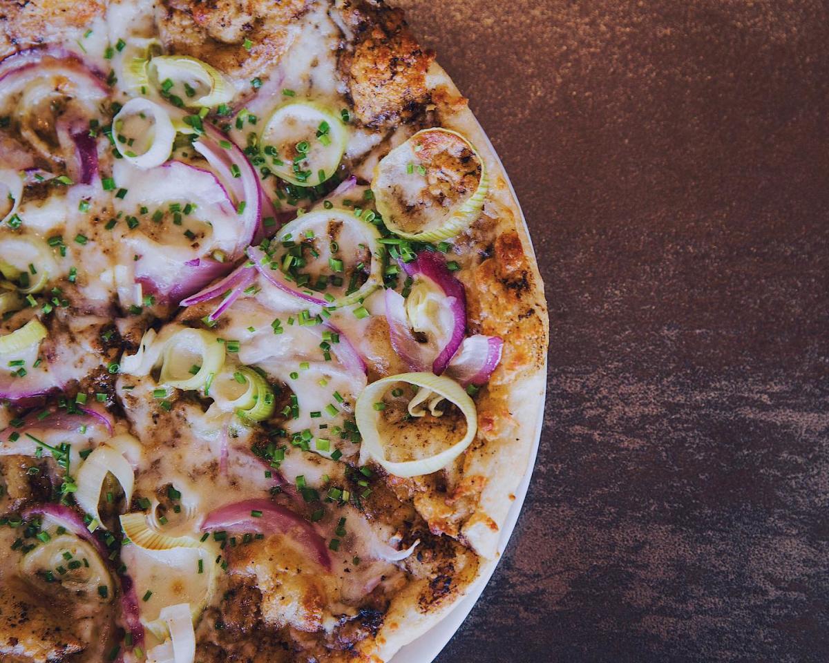 Close up of a wood fired pizza topped with sliced leeks, red onions, herbs and melted cheese on a browned crust.