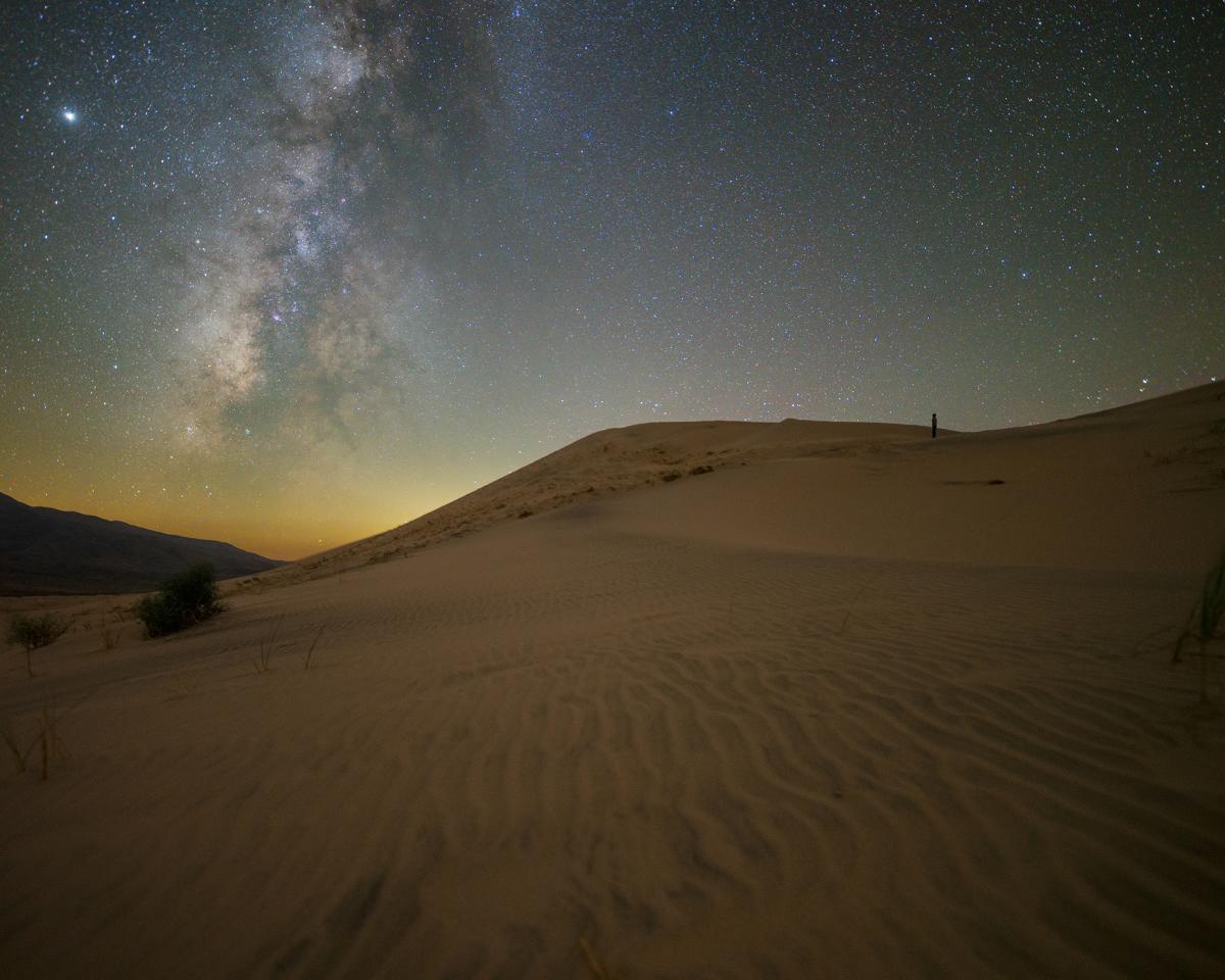 Photo of a person standing atop the Kelso Sand Dunes in Mojave National Preserve looking up to the stars and milky way. Photo by Jack Fusco.