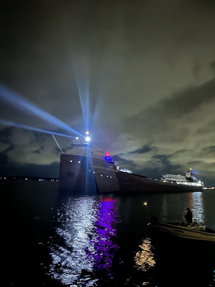 Nighttime view of a large ship illuminated by bright blue searchlight beams at a harbor, with reflections shimmering on the water's surface.