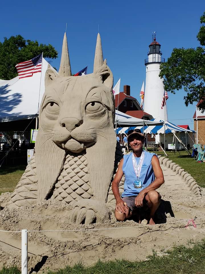 A man in a blue cutoff shirt wearing a medal around his neck kneels next to a sand sculpture of a cat-like figure with fangs, horns, and scales on the body.