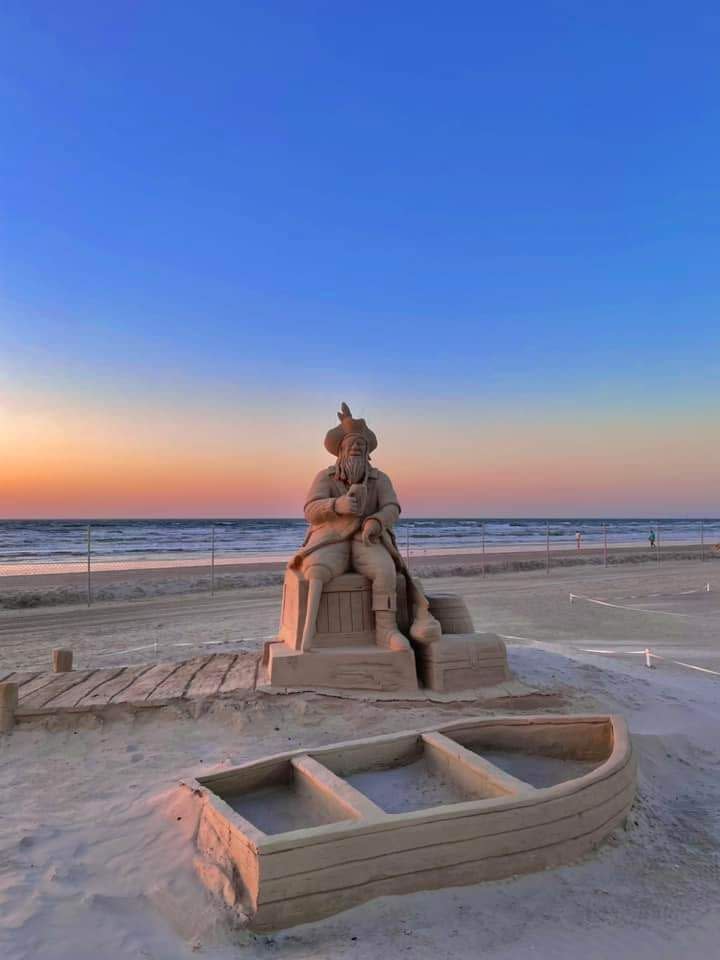 Sand sculpture on a beach at sunrise. The pirate is sitting drinking beer. In front of him is an empty life boat.