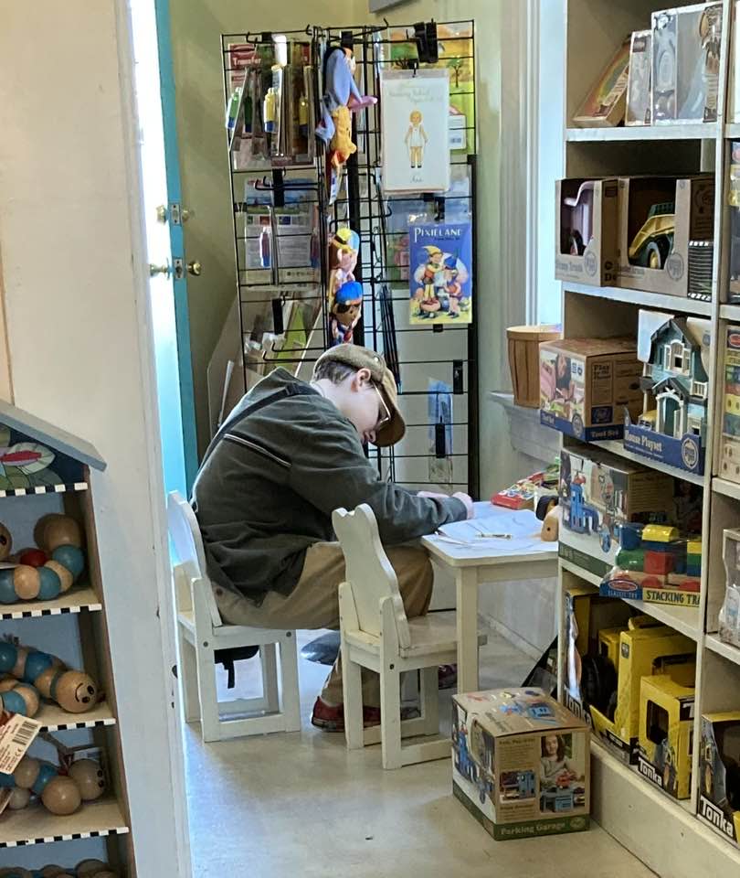 Image is of a young boy sitting at a small table and chair set coloring in a coloring book surrounded by shelves with toys.