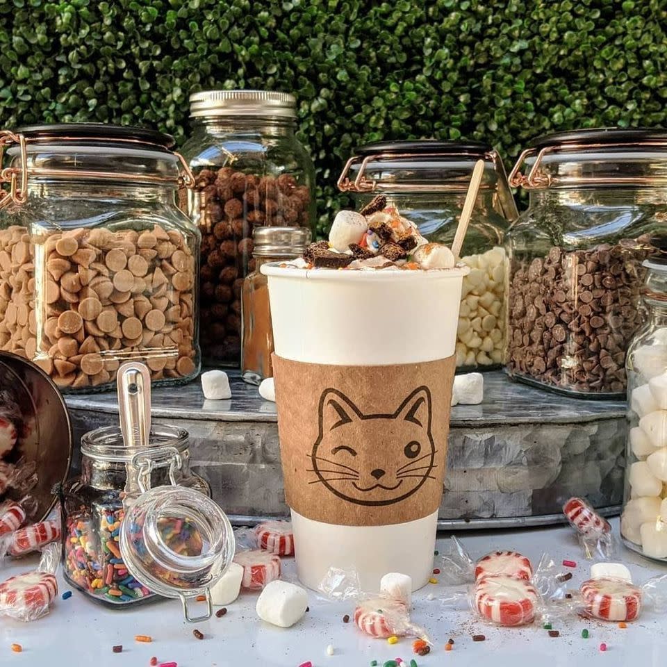 Image is of a white coffee cup to go with a cat image on the sleeve and candy toppings in glass jars surrounding the coffee.