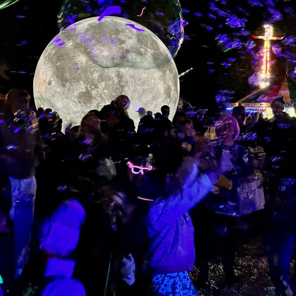 A large crowd gathers at night in front of a giant illuminated moon sculpture at an outdoor event. People are surrounded by glowing bubbles and wear light-up accessories like neon glasses and glow sticks. A carnival ride and colorful lights are visible in the background, creating a festive, dreamlike atmosphere.