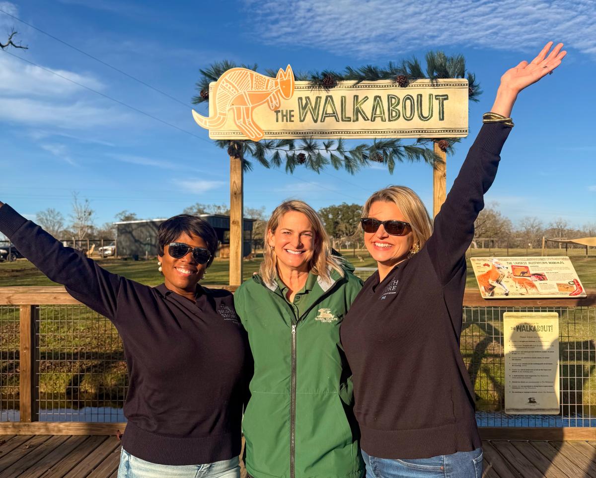 Christina Cooper, executive director of Global Wildlife Center, stands on a boardwalk between All The Waves co-hosts Zondra White Jones and Katie Guasco beneath a “The Walkabout” sign, all smiling with raised arms.