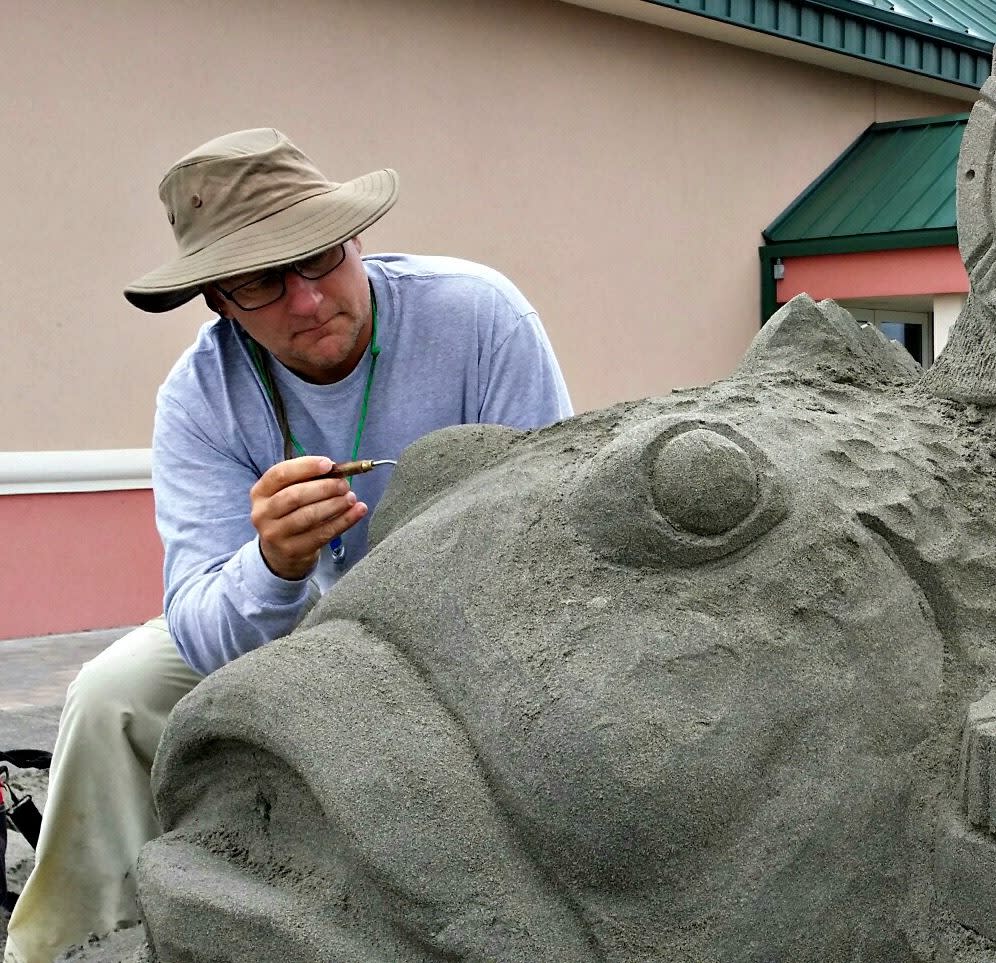 A man bends over a large sand sculpture of a fish with a paintbrush. He is wearing a bucket hat and focuses on his work.