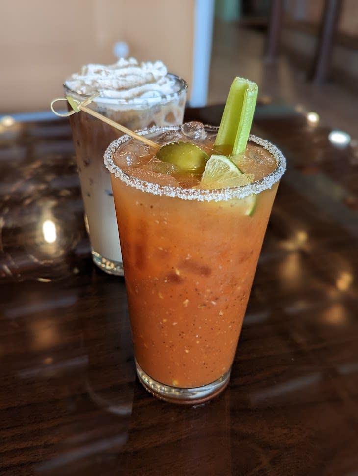 Image is of a bloody mary in the front with a celery stick, line and olive and in the background an iced coffee with whipped cream on top.