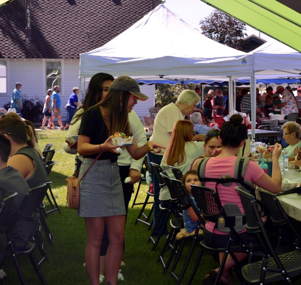Two young women, hands full with loaded baked potatoes, search for seating at packed picnic tables during Potato Day at the Centennial Village Museum