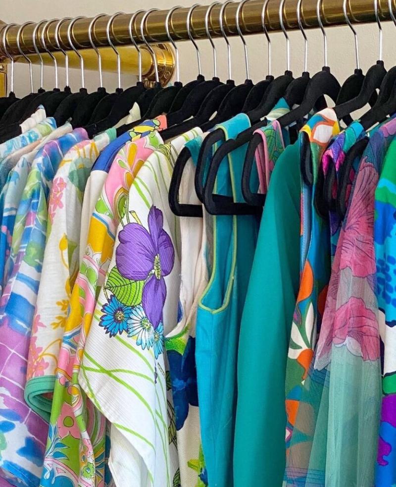 A variety of colorful garments hanging on black hangers from a gold-toned clothes rail, featuring bright floral patterns and solid colors.