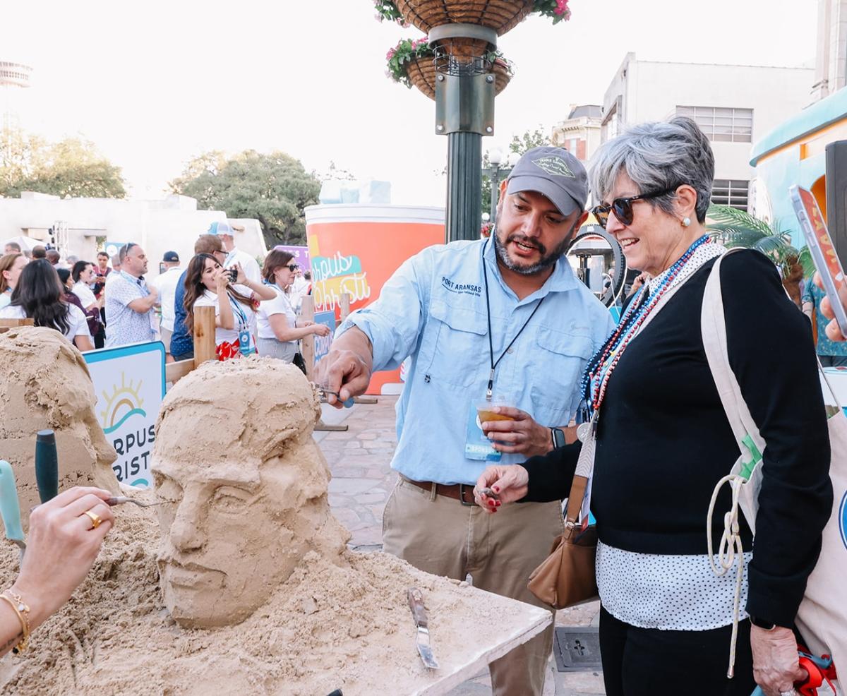 A man with a sand sculpting tool in a blue shirt shows a woman in a small sand sculpture how