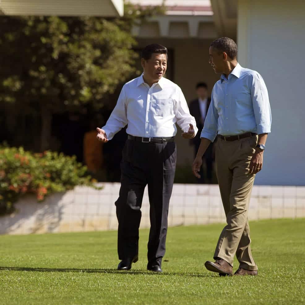 Two men walking and talking together on the grass with a building in the background. They are both dressed in business casual attire with shirts untucked and sleeves rolled up.