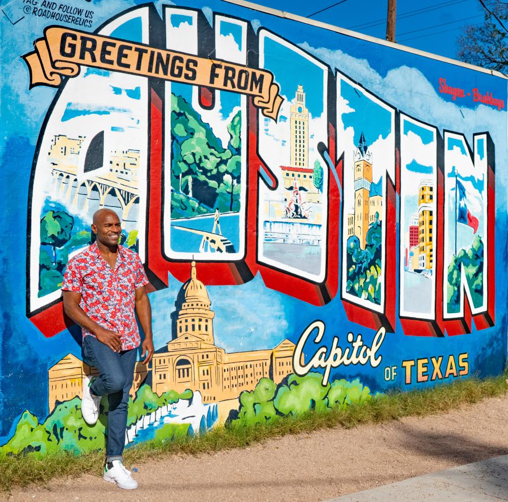 Man in patterned shirt leaning against the "Greetings from Austin" postcard mural.