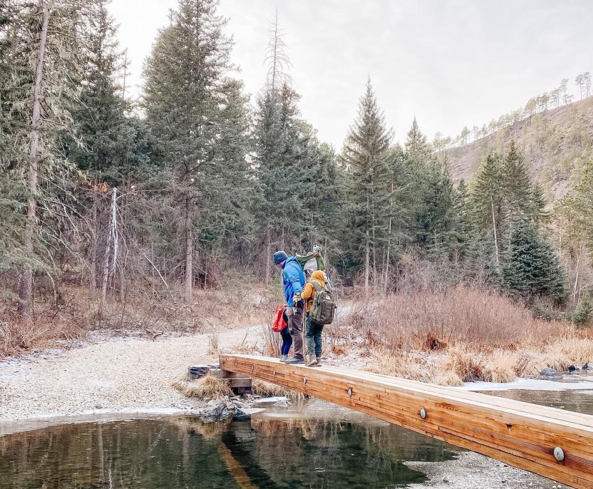 A couple is crossing a narrow wooden plank over a clear stream in a forest. Tall evergreen trees surround them, reflecting a calm, adventurous mood.