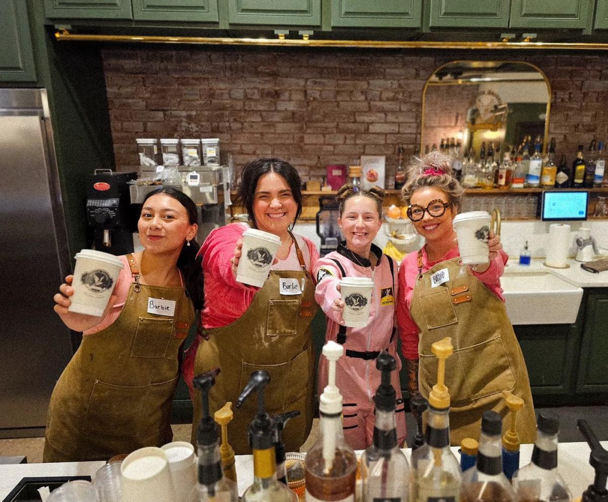 Four women employees at Greater Grounds Coffee Co. pose for a photo at the counter holding up coffee cups.