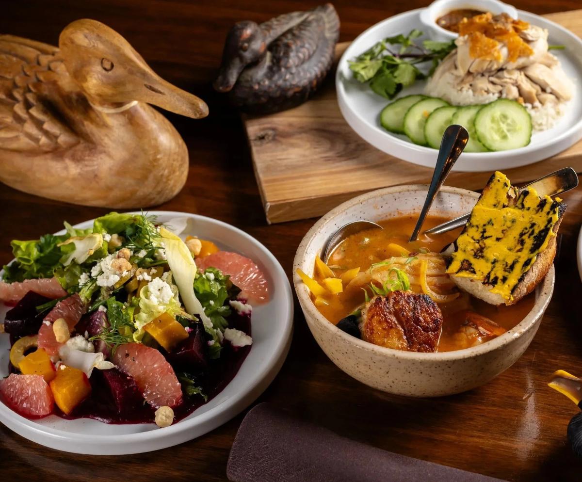 Spread of dishes at Odd Duck, including a citrus and beet salad, seafood stew with a seared scallop and grilled bread and a chicken and rice plate with cucumber slices.