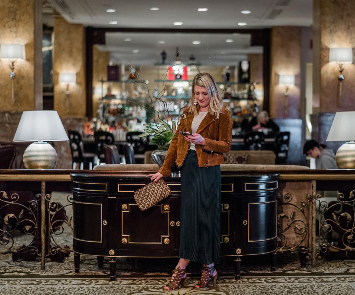 Interior photo in a hotel lobby lounge. A woman with blonde hair stands in front of a dark wood console table, looking at her phone and holding a patterned clutch. She is wearing a tan jacket, a long dark skirt, and heeled sandals. Warm lighting, decorative lamps, and a bar area with guests are visible in the background.