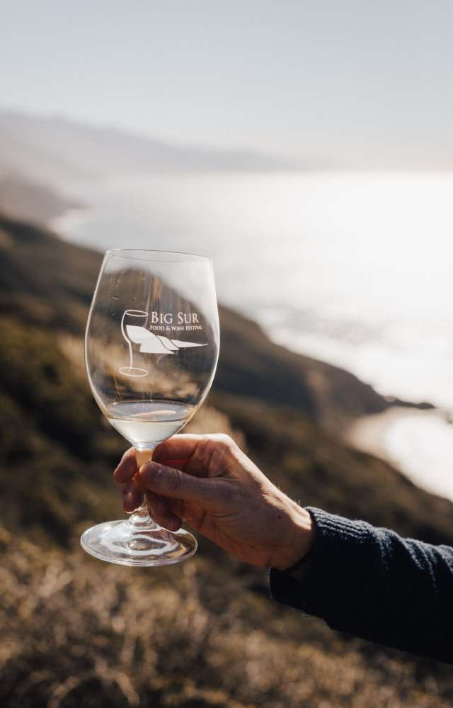 This is an image of a clear commemorative glass from Big Sur Food & Wine Festival held up against the dramatic coastline backdrop of Big Sur, California. The rolling mountains lead straight in to the ocean waters