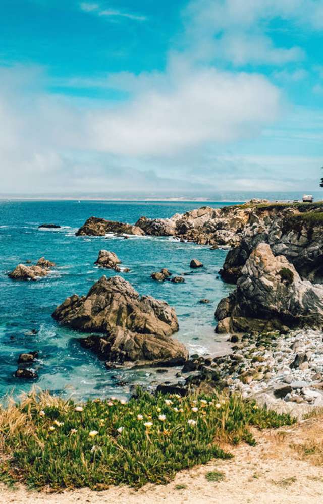 This is an image of the ocean view from the Monterey Bay Coastal Recreation Trail in Pacific Grove. The sandy trail is in the foreground with the rocky coastal shore of Pacific Grove and deep blue ocean in the background.