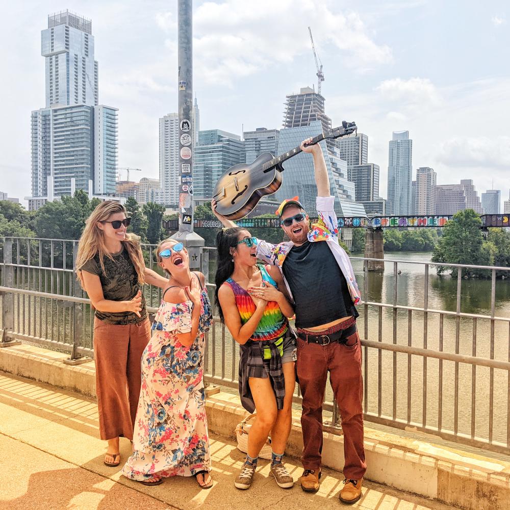 Four people posing on a bridge in front of Austin downtown skyline. Left to Right: woman grins at women beside her, woman places hand over heart and looks towards camera, woman places hands over her heart, man raises a guitar over his head