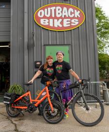Kate and Pete posing with two bikes in front of the Outback Bikes warehouse with the logo behind them.