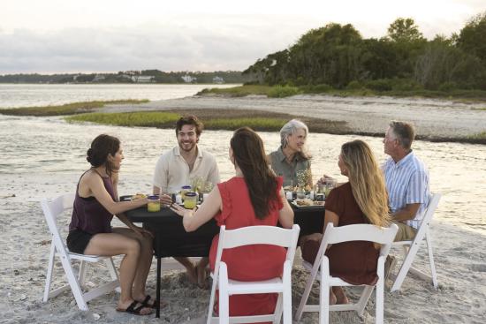 Group of men and women are sitting on white chairs at a white table in Wrightsville Beach enjoying a picnic dinner as the sun is setting with lime margaritas