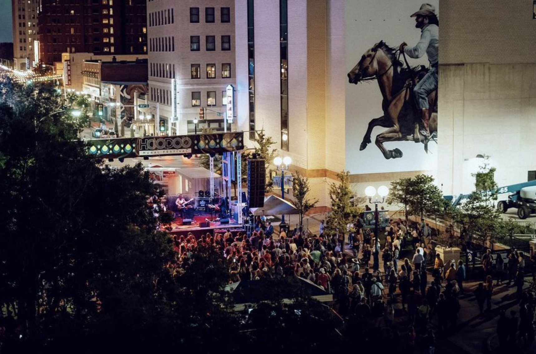 Photo of large audience in downtown amarillo with a mural featuring a man wearing a white shirt riding a brown horse and stage with purple lights