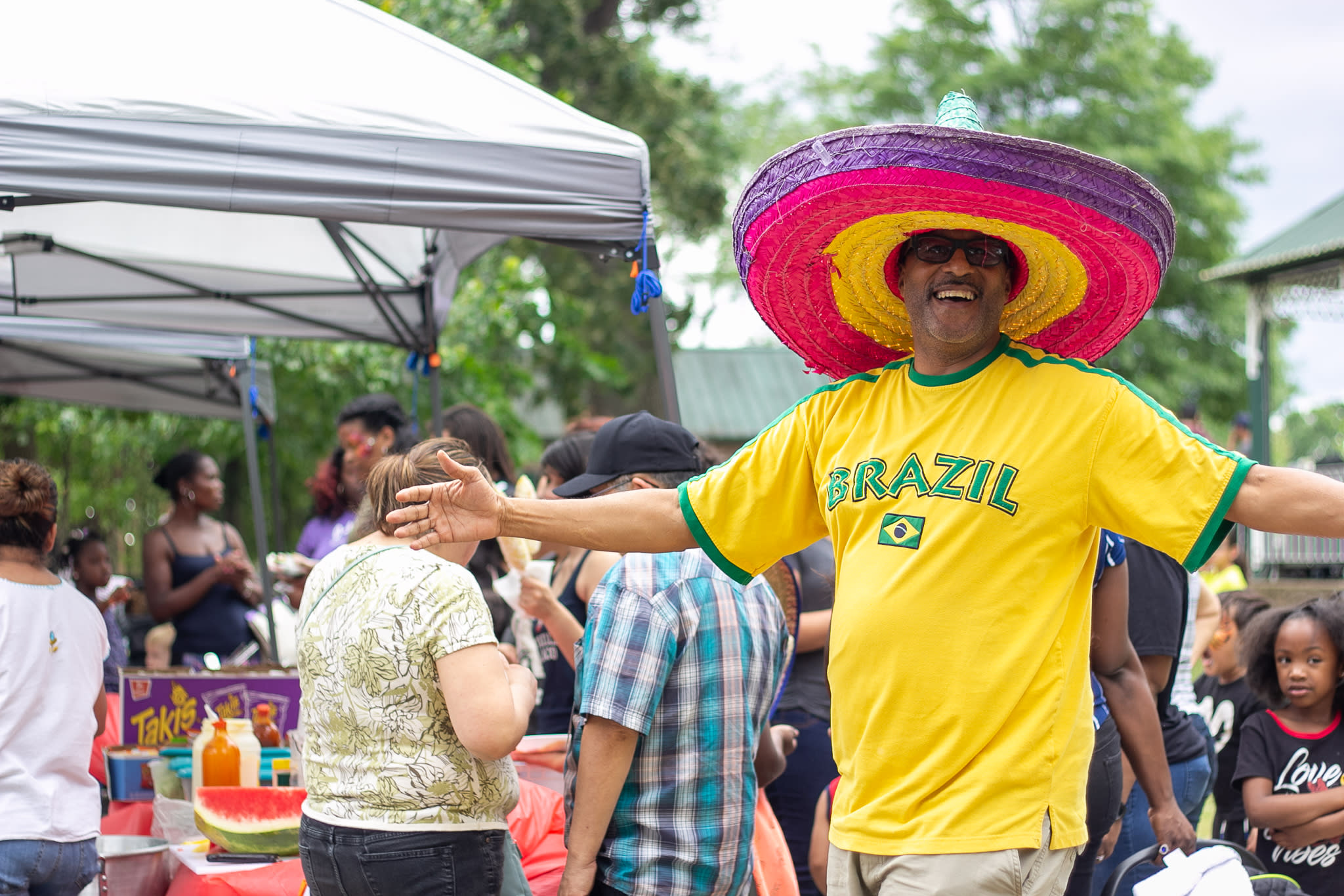 A man wearing a yellow Brazil shirt and pink, purple, and yellow sombrero poses with his arms open wide at La Fiesta.