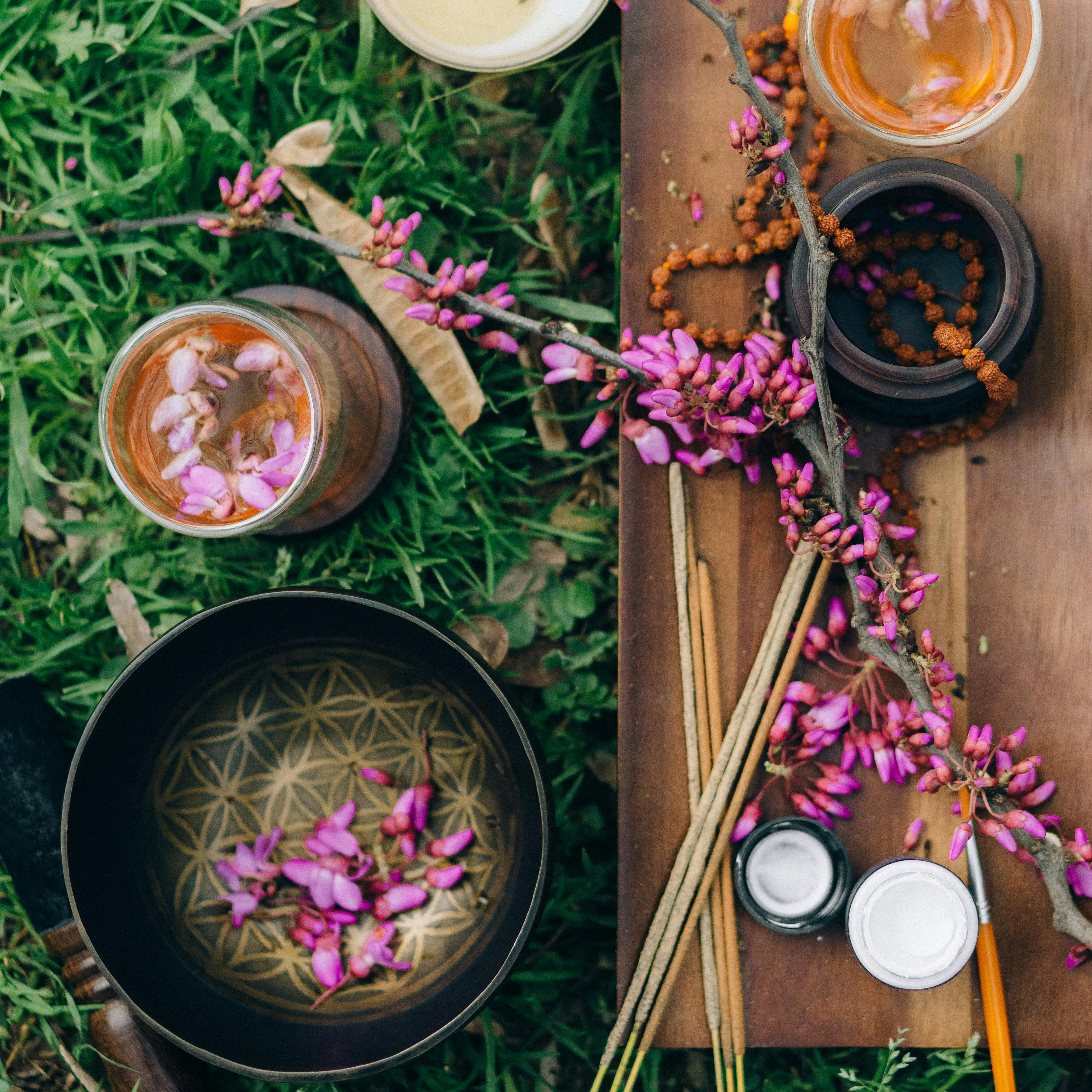 Herbal tea, pink blossoms, incense and botanical ingredients arranged outdoors on a wooden board.