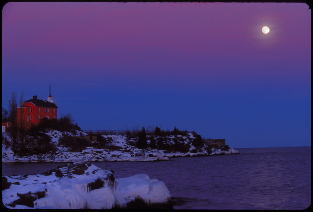 THE MARQUETTE LIGHTHOUSE WITH MOONRISE AND ICY SHORES IN WINTER IN MARQUETTE MICHIGAN.