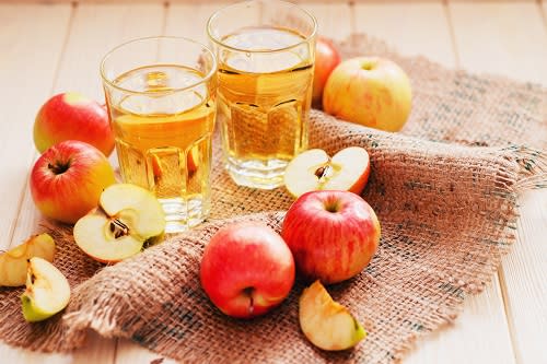 Two glasses of cider and a variety of apples - both whole and cut - sit on a burlap placemat on a wooden table