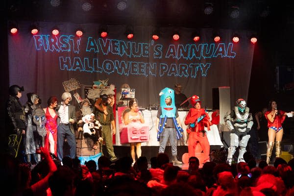 About a dozen people in costume stand on stage in front of a banner that says "First Avenue's Annual Halloween Party"