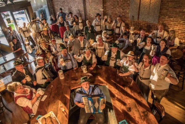 A room full of people dressed in traditional German attire play instruments, sing, and raise glasses of beer for a toast at Delano Oktoberfest